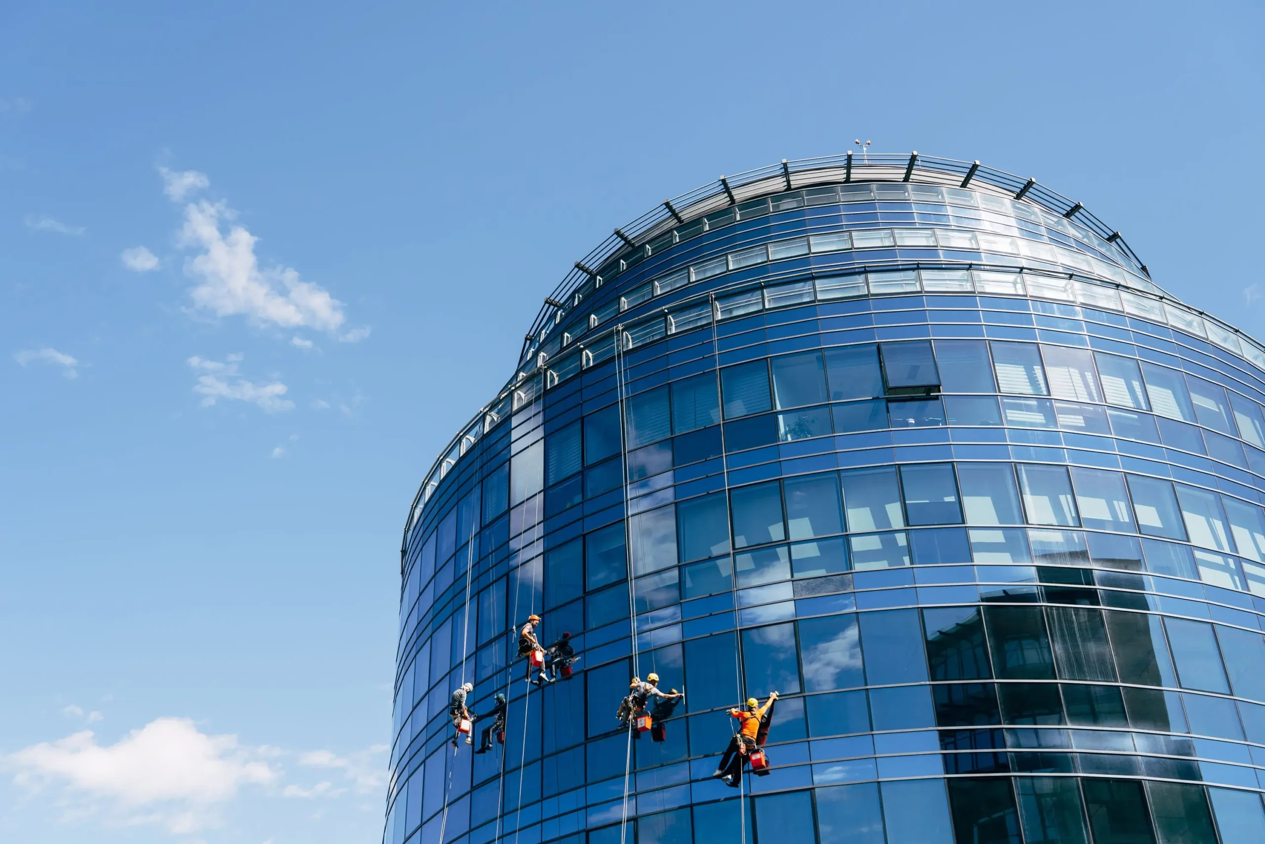 Dieses Bild zeigt ein modernes, hoch aufragendes Bürogebäude mit einer gebogenen Glasfassade. Mehrere Arbeiter, die Sicherheitsausrüstung tragen, sind an Seilen befestigt und reinigen die Glasflächen des Gebäudes. Der Himmel ist klar und blau, was eine helle und klare Umgebung für die Arbeit schafft. Die Szene zeigt deutlich die Höhe des Gebäudes und die Herausforderungen der Fassadenreinigung.