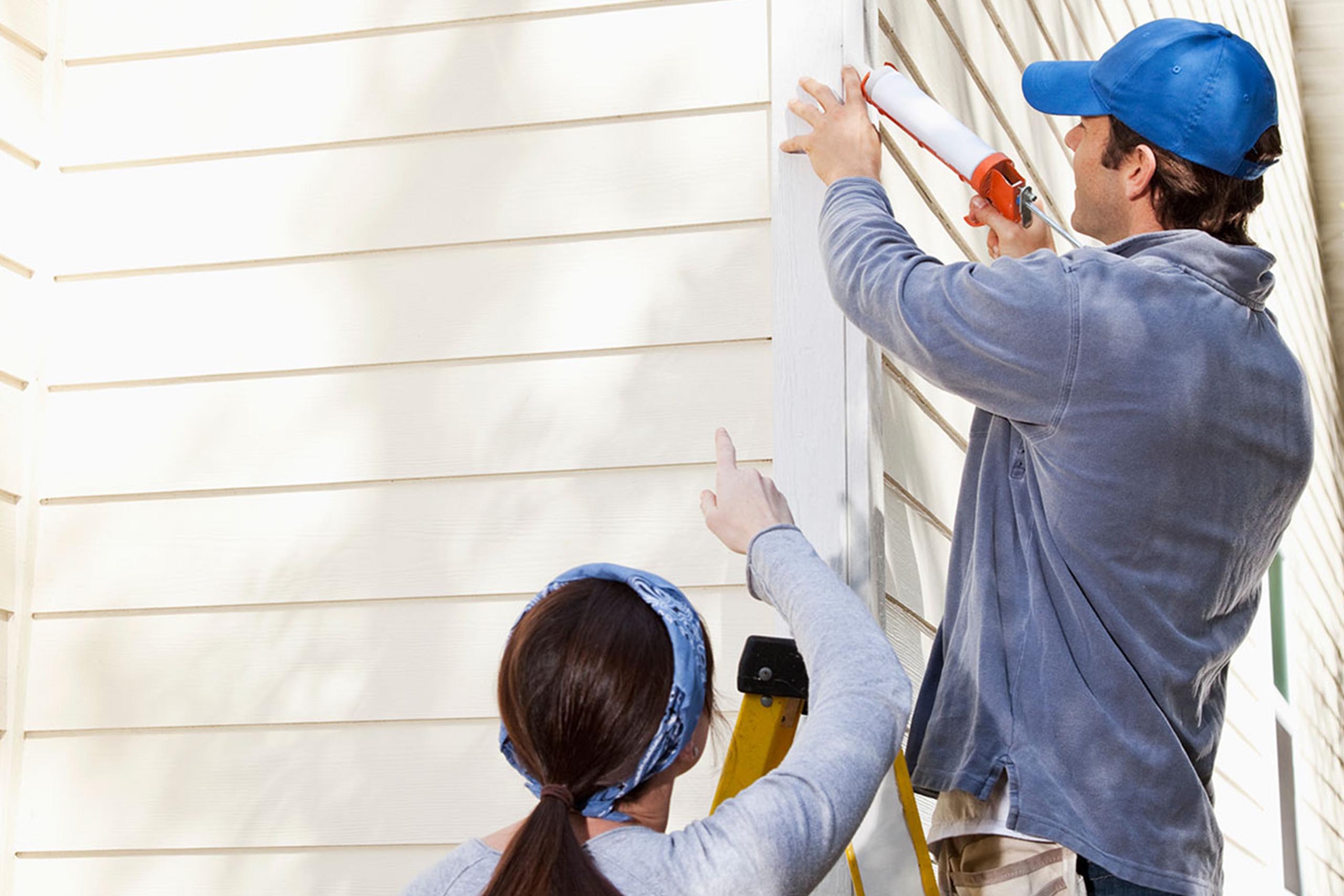 Dieses Bild zeigt zwei Personen, die Wartungsarbeiten an der Außenseite eines Gebäudes durchführen, was typisch für einen Hausmeisterservice ist. Eine Person steht auf einer Leiter und trägt eine blaue Kappe sowie eine graue Jacke. Sie verwendet eine Kartuschenpistole, um Dichtmittel auf einen Bereich der Gebäudefassade aufzutragen. Die zweite Person, die unten steht und auf die Arbeitsstelle zeigt, trägt ein graues Oberteil und ein blaues Kopftuch. Die Fassade des Gebäudes besteht aus hellen, horizontalen Paneelen. Diese Szene veranschaulicht typische Aufgaben eines Hausmeisterservices, wie das Abdichten und Instandhalten von Gebäuden.