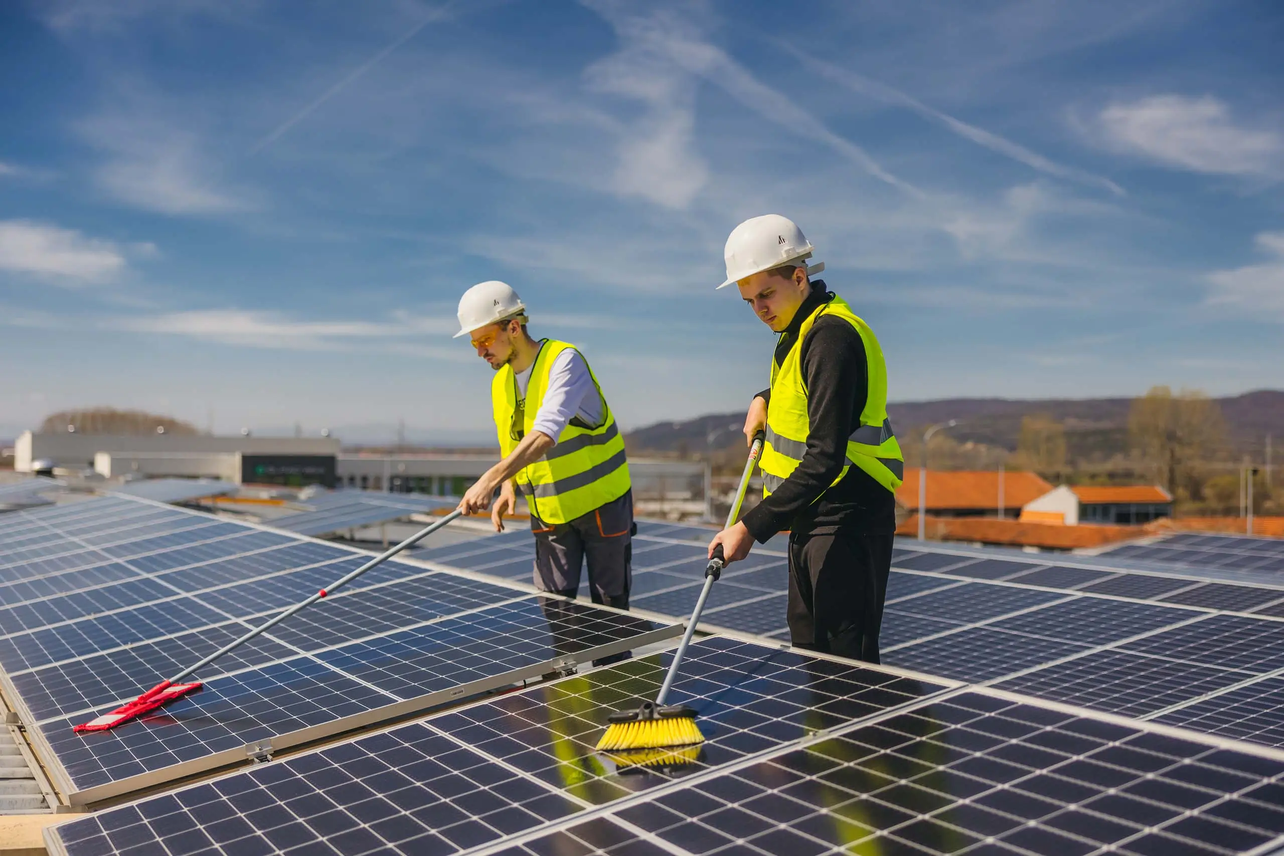 Dieses Bild zeigt zwei Personen, die Photovoltaikanlagen auf einem Dach reinigen. Beide tragen gelbe Warnwesten und weiße Schutzhelme, was auf die Arbeitssicherheit hinweist. Sie benutzen lange Bürsten, um die Solarmodule zu säubern. Der Himmel ist klar und blau, und im Hintergrund sind Berge und weitere Gebäude zu sehen. Diese Szene veranschaulicht die Reinigung und Wartung von Solaranlagen, um deren Effizienz und Langlebigkeit sicherzustellen.