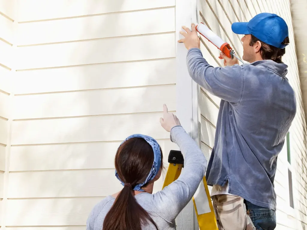 Dieses Bild zeigt zwei Personen, die Wartungsarbeiten an der Außenseite eines Gebäudes durchführen, was typisch für einen Hausmeisterservice ist. Eine Person steht auf einer Leiter und trägt eine blaue Kappe sowie eine graue Jacke. Sie verwendet eine Kartuschenpistole, um Dichtmittel auf einen Bereich der Gebäudefassade aufzutragen. Die zweite Person, die unten steht und auf die Arbeitsstelle zeigt, trägt ein graues Oberteil und ein blaues Kopftuch. Die Fassade des Gebäudes besteht aus hellen, horizontalen Paneelen. Diese Szene veranschaulicht typische Aufgaben eines Hausmeisterservices, wie das Abdichten und Instandhalten von Gebäuden.