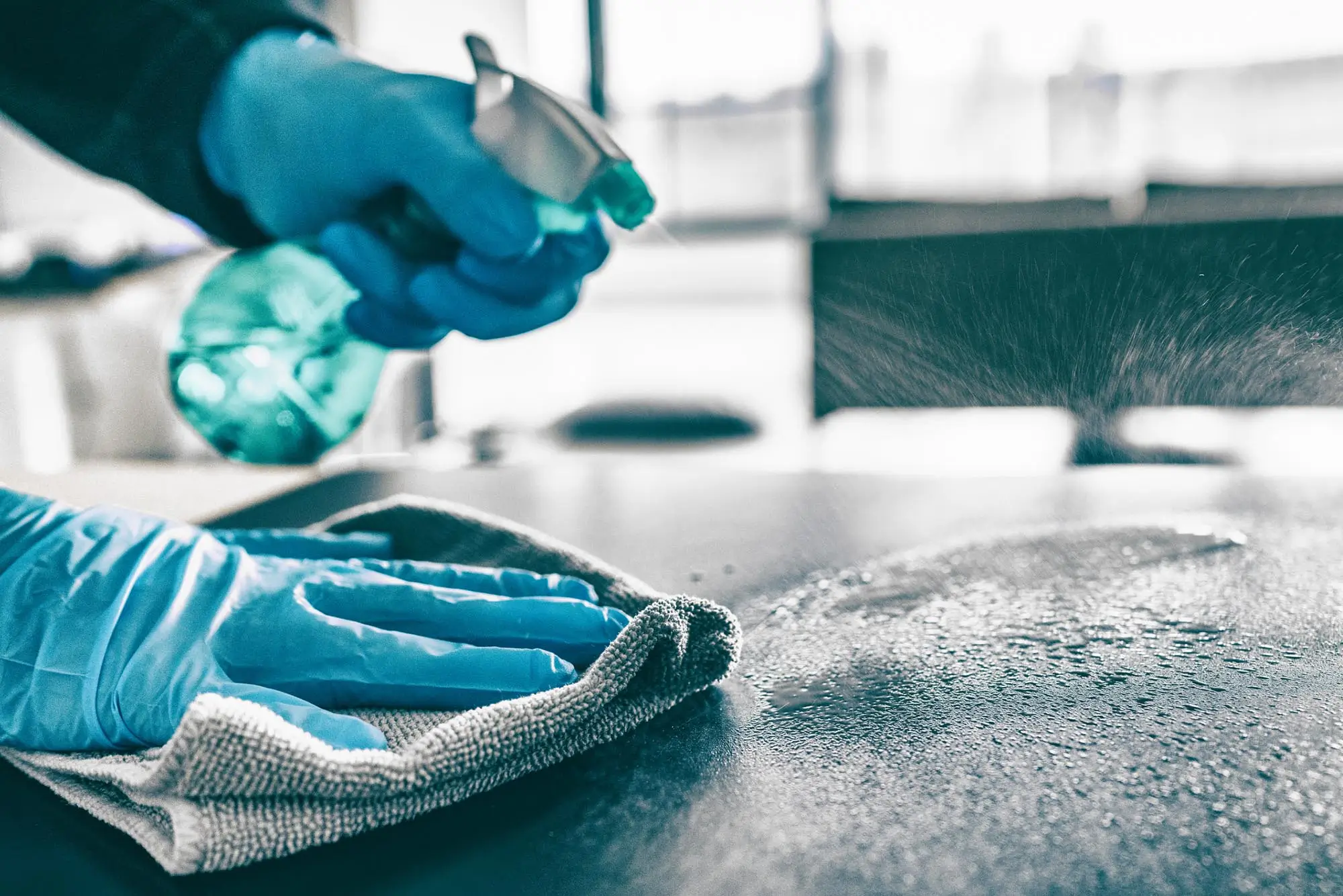 Dieses Bild zeigt eine Nahaufnahme einer Hand, die mit blauen Gummihandschuhen eine Oberfläche reinigt. Die Person hält eine Sprühflasche mit grünem Reinigungsmittel und sprüht es auf die Oberfläche, während sie mit einem Tuch die Flüssigkeit verteilt und abwischt. Die Szene symbolisiert Tiefenreinigung und Desinfektion, bei der gründlich gereinigt und desinfiziert wird, um Bakterien, Viren und Schmutz zu entfernen und eine hygienische Umgebung zu gewährleisten.