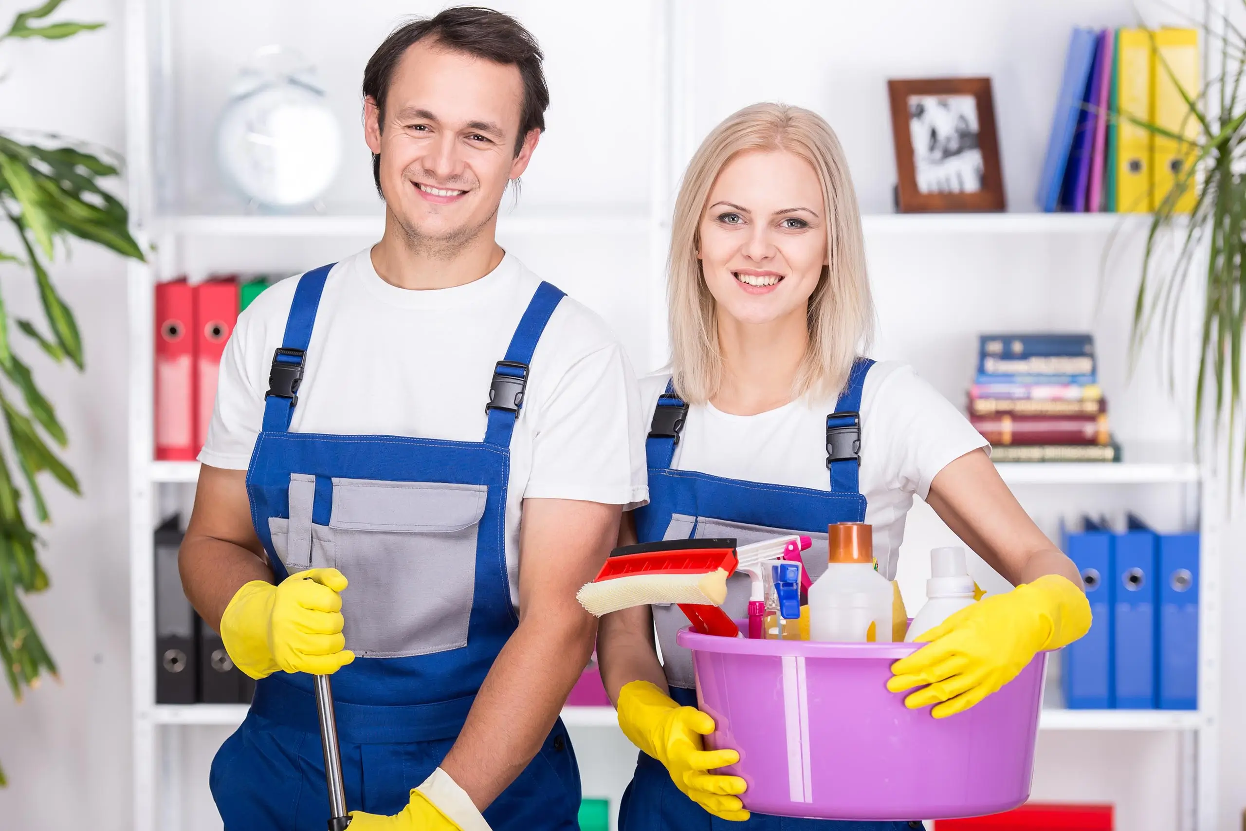 Dieses Bild zeigt zwei Personen in Arbeitskleidung, die für Reinigungsarbeiten bereitstehen. Beide tragen blaue Latzhosen, weiße T-Shirts und gelbe Gummihandschuhe. Die Person auf der linken Seite hält einen Wischmopp, während die Person auf der rechten Seite einen violetten Eimer mit verschiedenen Reinigungsutensilien, wie Bürsten und Reinigungsmitteln, trägt. Beide lächeln freundlich. Im Hintergrund sind Regale mit Büchern und Ordnern zu sehen. Das Bild steht symbolisch für umweltfreundliche Reinigung, was durch die freundliche und professionelle Ausstrahlung der Personen sowie die Präsenz von Reinigungsmitteln angedeutet wird, die darauf hinweisen, dass die Reinigungsarbeiten umweltbewusst durchgeführt werden.