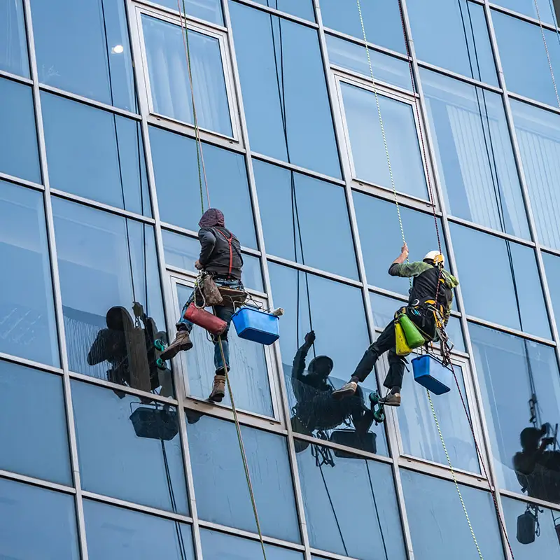 Dieses Bild zeigt zwei Fensterreiniger, die an Seilen befestigt sind und an der Glasfassade eines hohen Gebäudes arbeiten. Sie sind mit Sicherheitsgurten, Eimern und Reinigungswerkzeugen ausgestattet und reinigen die großen Fenster des Gebäudes. Die Reiniger tragen Schutzkleidung und verwenden Techniken, die Präzision und Sorgfalt erfordern, um sicherzustellen, dass die Fenster makellos sauber sind. Dieses Bild steht für die Reinigung von Hochhausfenstern, bei der die Außenfenster hoher Gebäude gereinigt werden, was oft den Einsatz spezieller Ausrüstung und Sicherheitsmaßnahmen erfordert.