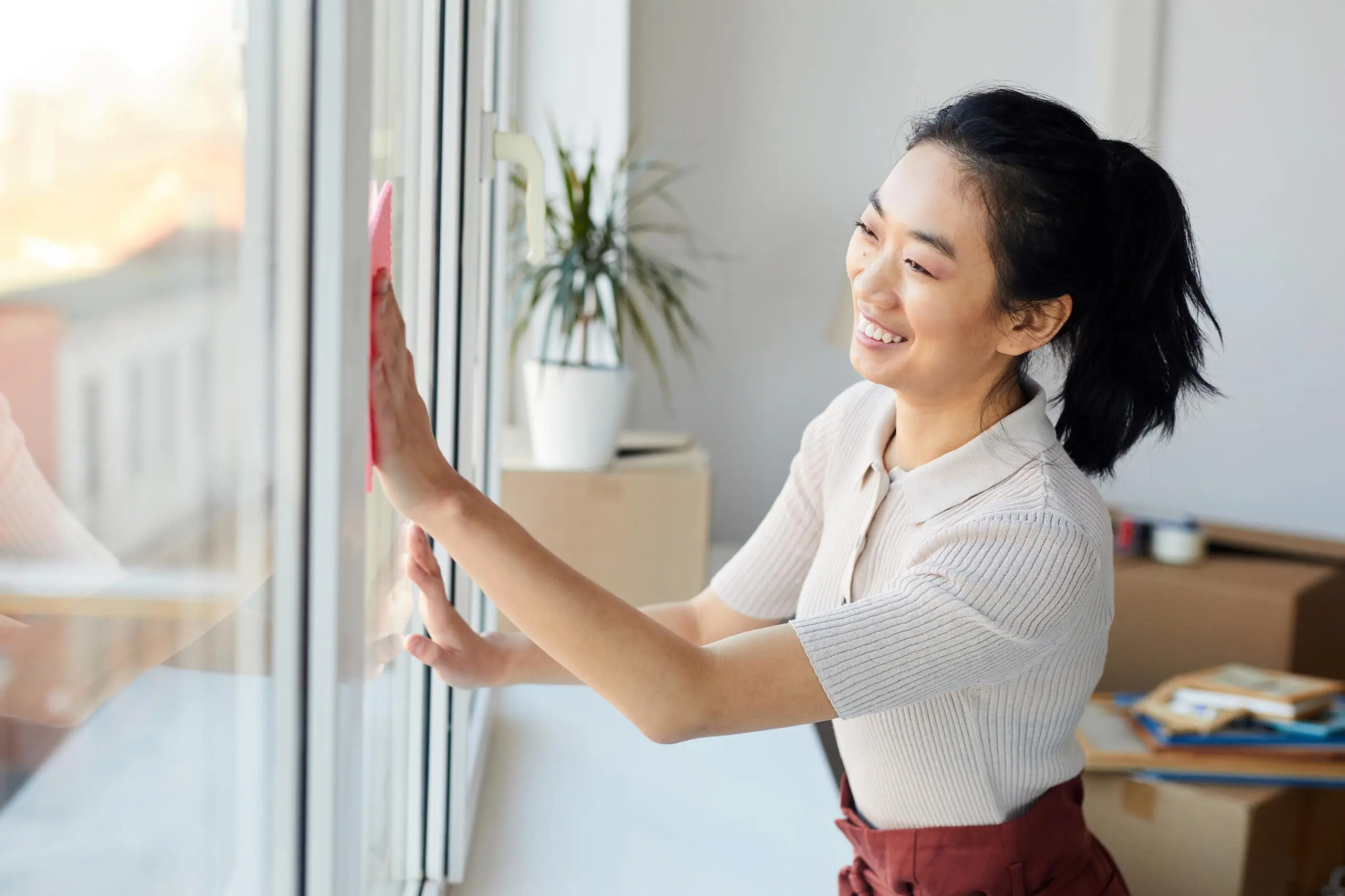 Dieses Bild zeigt eine Frau, die ein Fenster reinigt. Sie lächelt und verwendet ein rosa Reinigungstuch, um die Glasfläche zu säubern. Im Hintergrund ist ein heller Raum mit einigen Pflanzen und Gegenständen zu sehen. Die Szene wirkt freundlich und einladend. Dieses Bild steht für die Rahmenreinigung, bei der nicht nur die Glasfläche, sondern auch die Fensterrahmen gründlich gereinigt werden, um eine umfassende Sauberkeit zu gewährleisten.