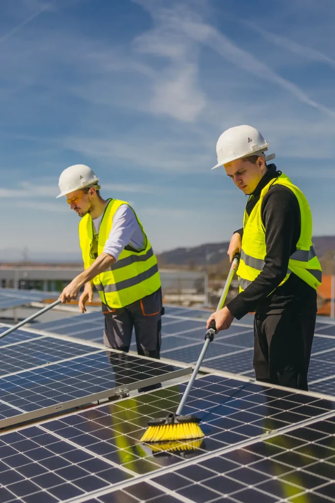 Dieses Bild zeigt zwei Personen, die Photovoltaikanlagen auf einem Dach reinigen. Beide tragen gelbe Warnwesten und weiße Schutzhelme, was auf die Arbeitssicherheit hinweist. Sie benutzen lange Bürsten, um die Solarmodule zu säubern. Der Himmel ist klar und blau, und im Hintergrund sind Berge und weitere Gebäude zu sehen. Diese Szene veranschaulicht die Reinigung und Wartung von Solaranlagen, um deren Effizienz und Langlebigkeit sicherzustellen.