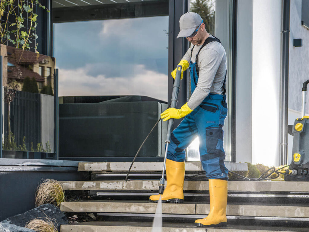Bild zeigt eine Person, die eine Treppe im Freien mit einem Hochdruckreiniger säubert. Die Person trägt eine blaue Latzhose, gelbe Gummistiefel und gelbe Gummihandschuhe sowie eine graue Kappe. Sie richtet den Wasserstrahl des Hochdruckreinigers auf die Treppenstufen, um diese zu reinigen. Im Hintergrund ist ein modernes Gebäude mit großen Glasfenstern zu sehen.