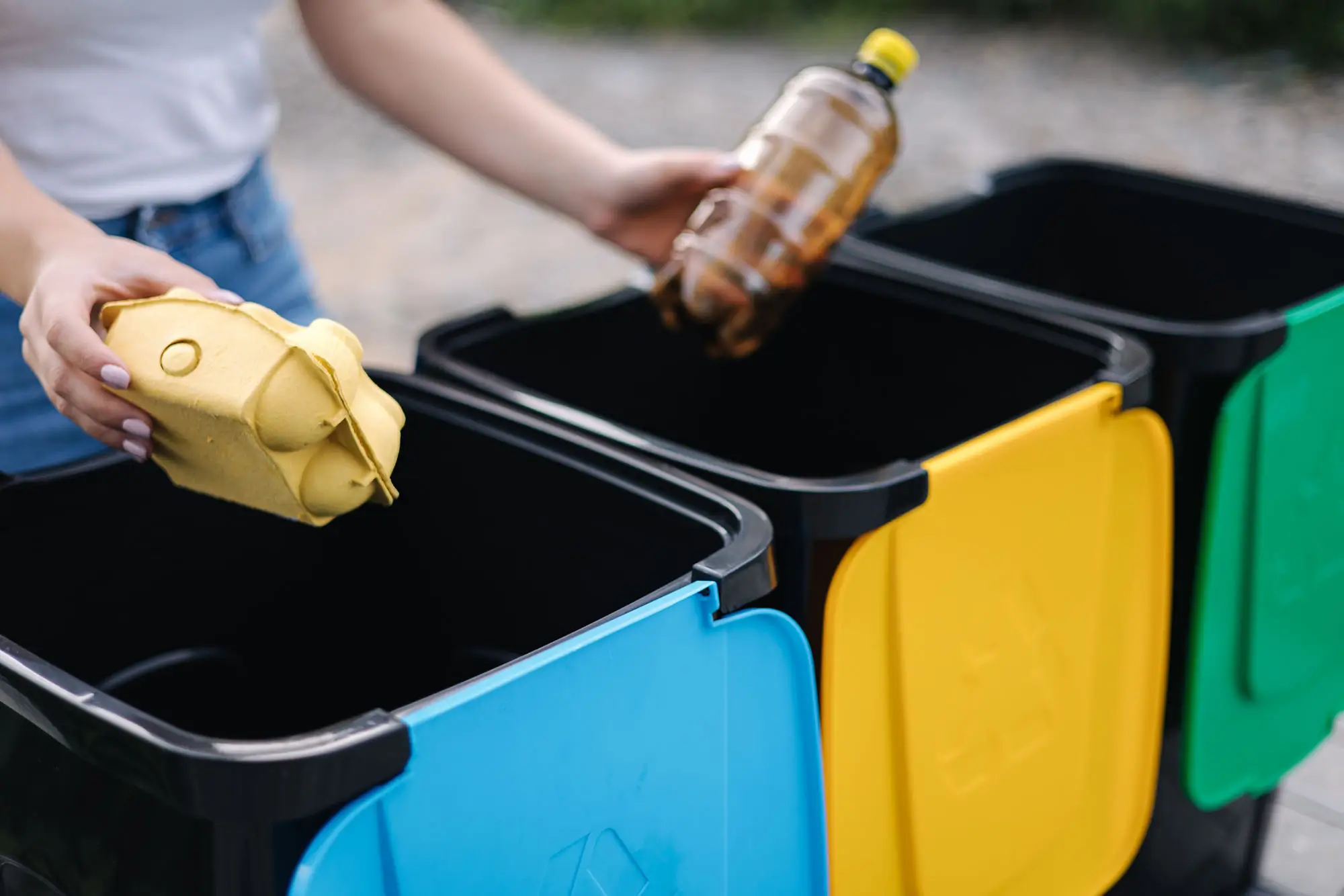 Dieses Bild zeigt eine Person, die Müll sortiert, indem sie Gegenstände in farblich gekennzeichnete Abfallbehälter legt. Die Person hält in einer Hand einen gelben Eierkarton und in der anderen eine leere Plastikflasche. Die Müllbehälter sind in verschiedenen Farben gekennzeichnet, was auf die Trennung von Abfall für Recyclingzwecke hinweist. Diese Szene symbolisiert Abfallentsorgung und Recycling, bei der Materialien getrennt und recycelt werden, um Umweltbelastungen zu reduzieren und Ressourcen zu schonen.