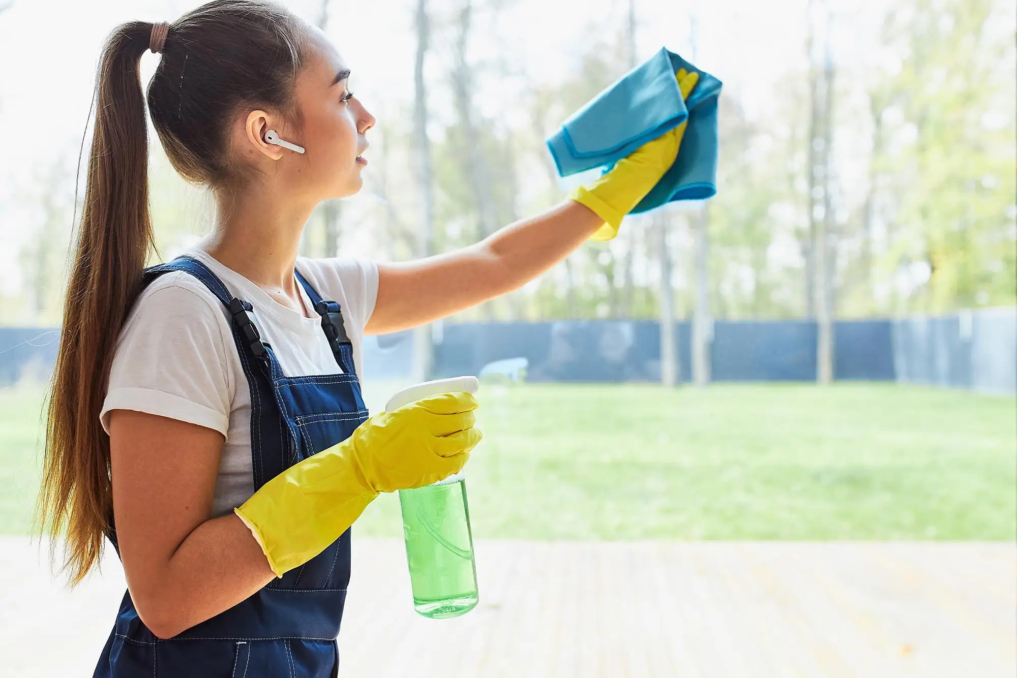 Dieses Bild zeigt eine junge Frau, die mit gelben Gummihandschuhen eine Glasoberfläche reinigt. Sie trägt eine blaue Latzhose und ein weißes T-Shirt. In einer Hand hält sie ein Reinigungstuch, mit dem sie das Glas wischt, und in der anderen Hand eine Sprühflasche mit grünem Reinigungsmittel. Im Hintergrund ist ein grüner Außenbereich zu sehen. Diese Szene steht für wöchentliche und monatliche Reinigungsdienste, bei denen regelmäßig geplante Reinigungsarbeiten durchgeführt werden, um die Sauberkeit und Pflege von Räumen zu gewährleisten.