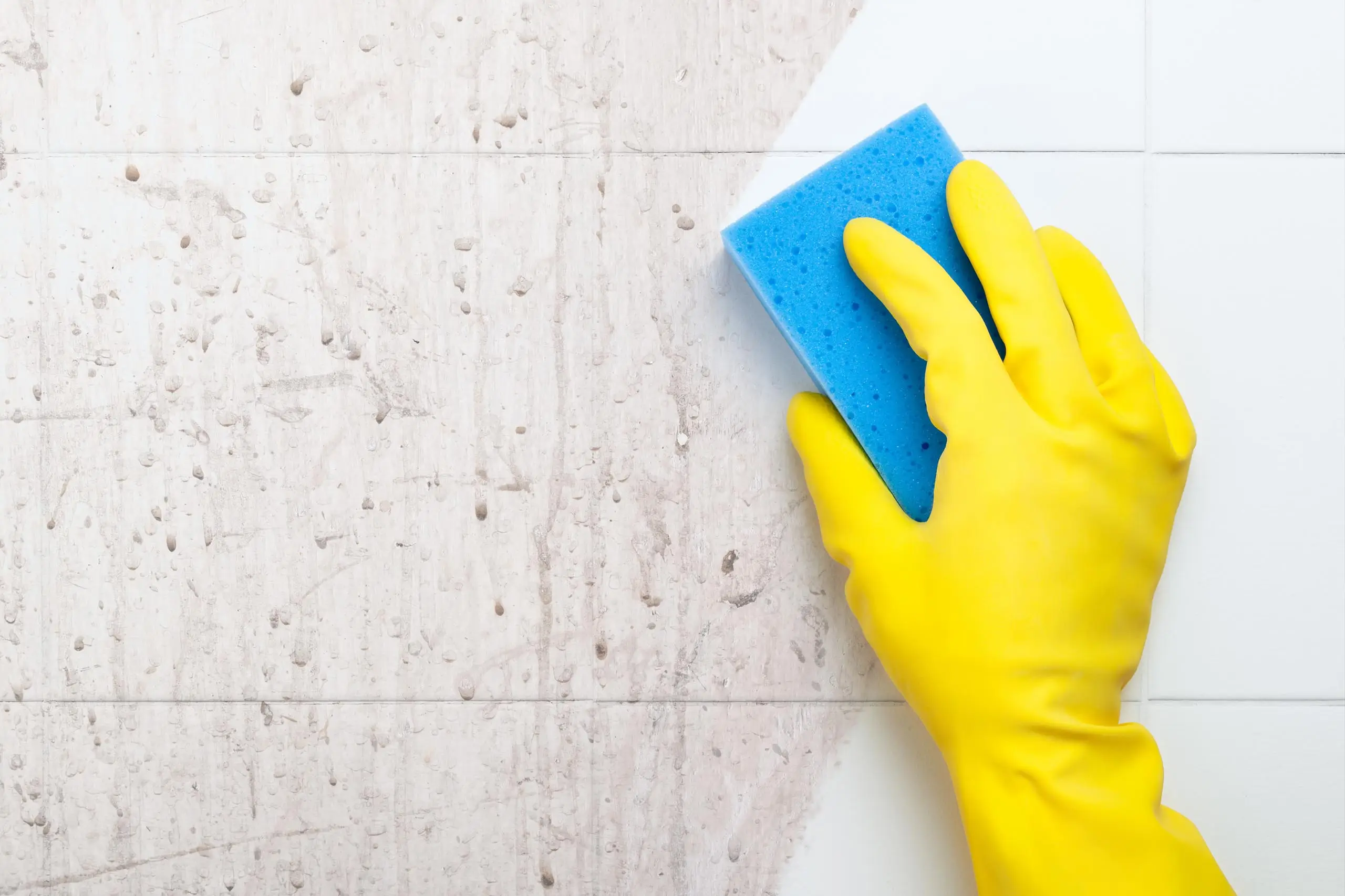 Dieses Bild zeigt eine Hand in einem gelben Gummihandschuh, die mit einem blauen Schwamm eine stark verschmutzte Oberfläche reinigt. Die Oberfläche ist mit hartnäckigen Flecken und Schmutz bedeckt, während ein Teil bereits sauber ist, wo der Schwamm verwendet wurde. Dieses Bild symbolisiert die Entfernung von hartnäckigen Verschmutzungen, bei der gründliche Reinigungsarbeiten durchgeführt werden, um stark verschmutzte Oberflächen wieder sauber und ansprechend zu machen.